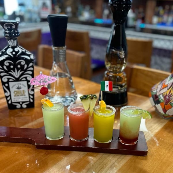 Four colorful margarita shots with garnishes on a wooden paddle, with tequila bottles in the background.