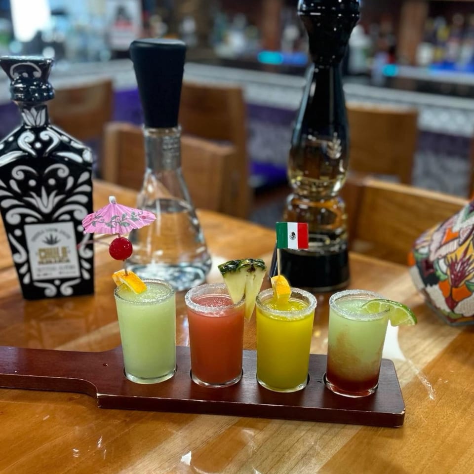 Four colorful margarita shots with garnishes on a wooden paddle, with tequila bottles in the background.