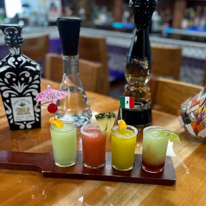 Four colorful margarita shots with garnishes on a wooden paddle, with tequila bottles in the background.