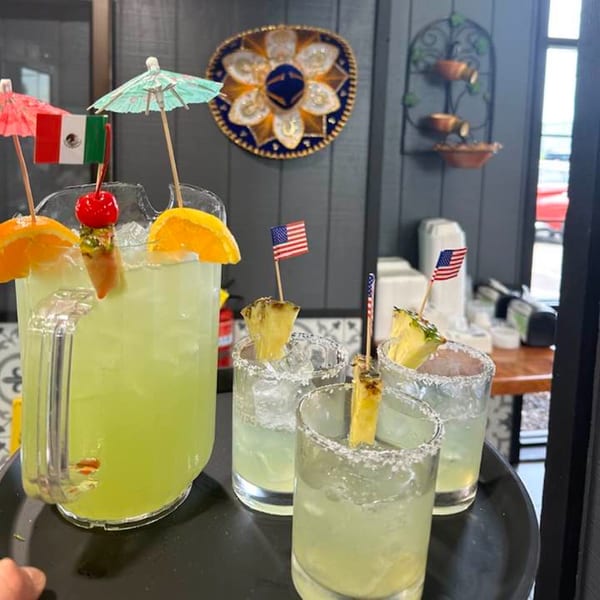 A pitcher of margaritas with orange slices and a cherry, alongside three glasses with pineapple and flags.