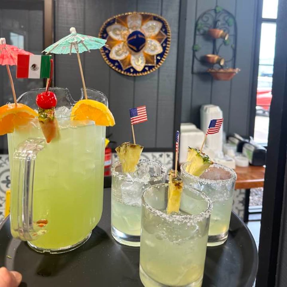 A pitcher of margaritas with orange slices and a cherry, alongside three glasses with pineapple and flags.