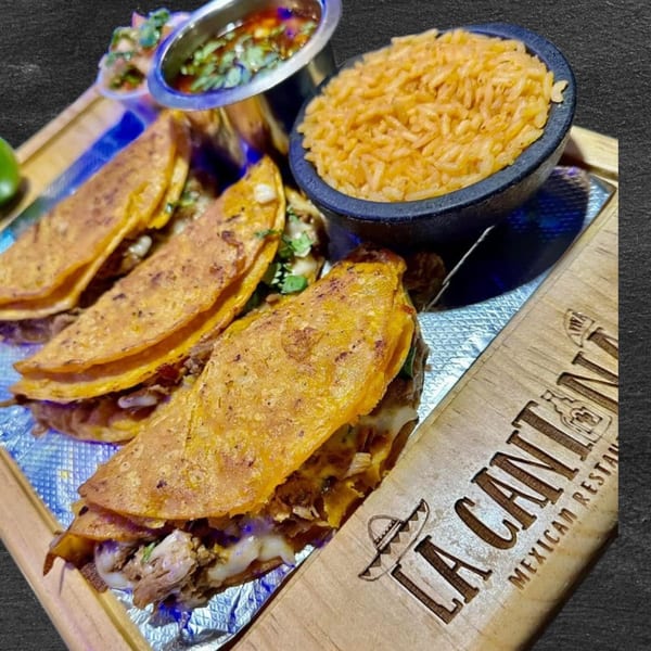 Three birria tacos with melted cheese, a side of rice, and consome on a wooden tray.