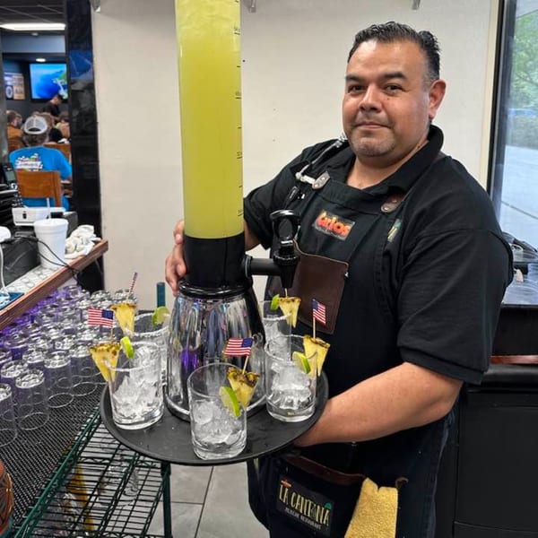 A La Cantina staff member holds a large margarita dispenser and a tray of garnished glasses.